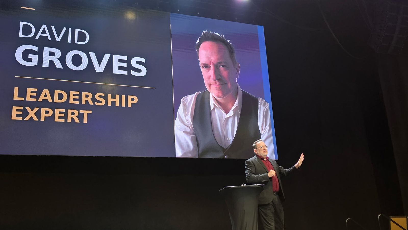 Dave Groves speaking on stage under a screen reading David Groves, Leadership Expert